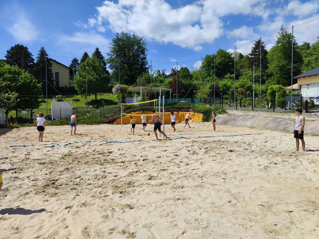 Eine Gruppe von Menschen spielt Beachvolleyball auf einem sandigen Feld mit Netz und Zaun. Bäume und ein Haus sind im Hintergrund.