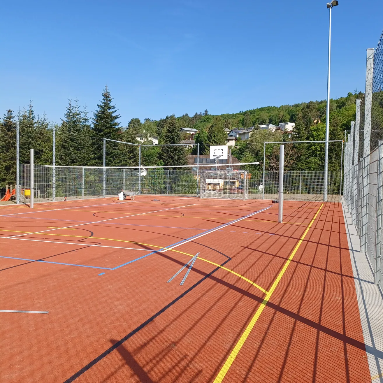 Ein Außensportplatz mit rotem Boden und blauen Markierungen. Rechts steht ein Basketballkorb, dahinter ein Volleyballnetz. An den Seiten befinden sich Zäune und im Hintergrund Bäume.