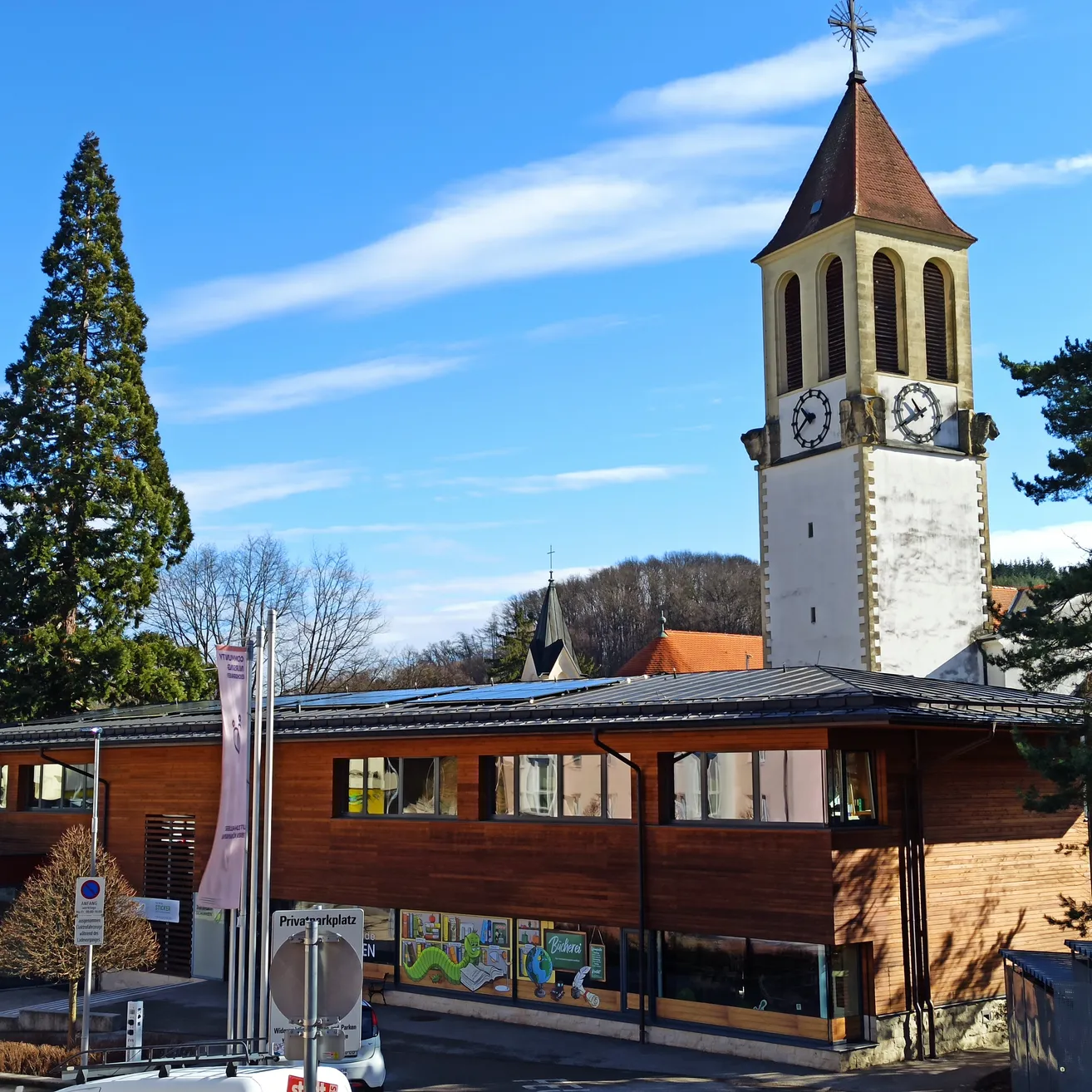 Ein hoher Glockenturm steht neben einem modernen Gebäude mit flachem Dach und Solarmodulen, unter einem blauen Himmel mit verstreuten Wolken.