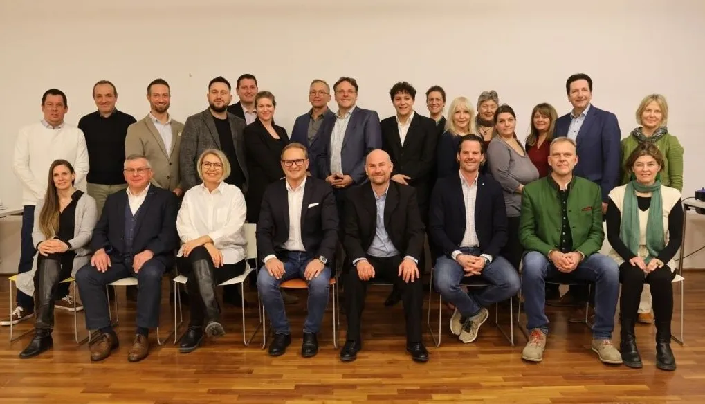 Eine Gruppe von Menschen, wahrscheinlich ein Team, posiert lächelnd für ein Foto. Sie sitzen auf Stühlen und stehen vor einer weißen Wand, gekleidet in formelle Kleidung.