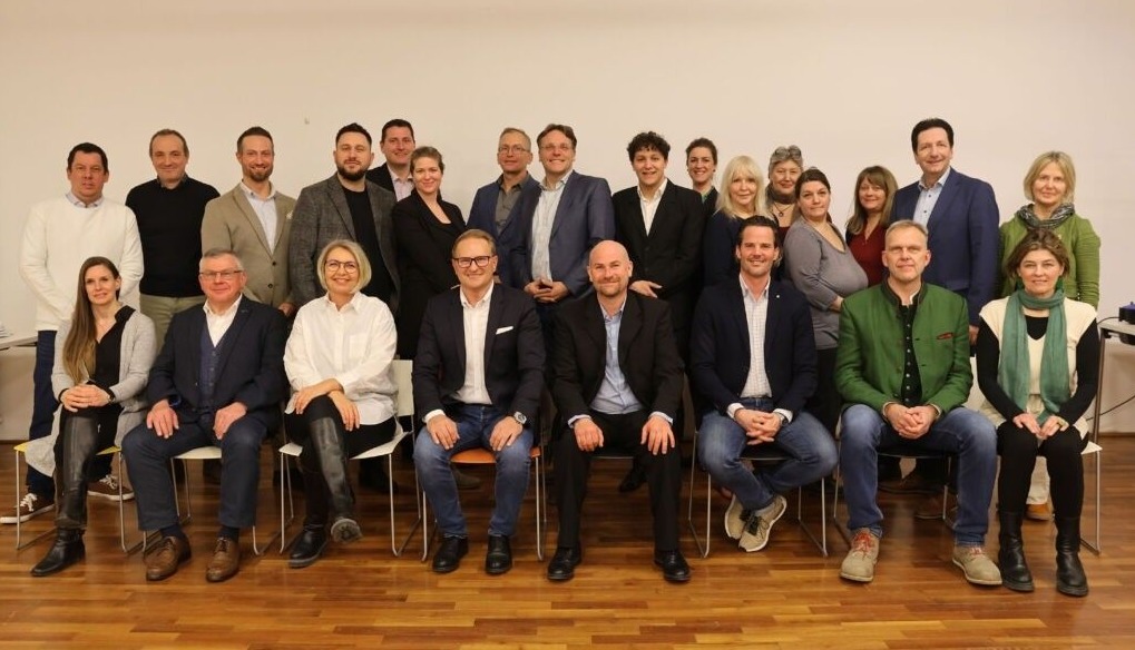 Eine Gruppe von Menschen, wahrscheinlich ein Team, posiert lächelnd für ein Foto. Sie sitzen auf Stühlen und stehen vor einer weißen Wand, gekleidet in formelle Kleidung.