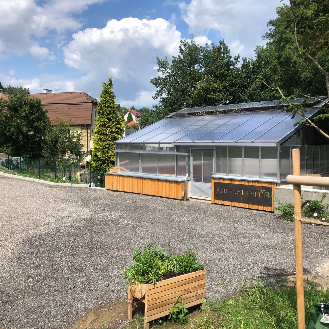 Ein großes Gewächshaus mit Solarpanelen auf dem Dach und einem Holzzaun befindet sich auf einer Kiesstraße. Ein hölzernes Schild steht neben dem Gewächshaus. Ein hölzerner Pflanzkasten mit Pflanzen steht vor dem Gewächshaus. Bäume und Häuser befinden sich hinter dem Gewächshaus.