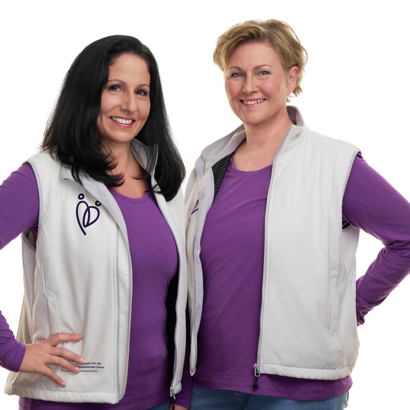 Zwei Frauen stehen eng beieinander und tragen beide eine weiße Weste mit einem lilafarbenen Shirt darunter. Sie lächeln beide und scheinen für ein Foto zu posieren.