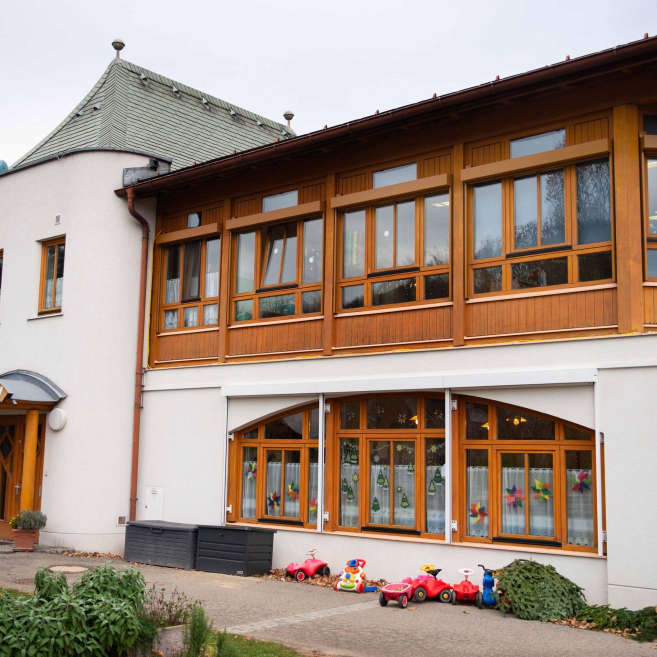 Ein weißes Gebäude mit Holzfassaden und Rundbogenfenstern. Das Gebäude ist mit Weihnachtsdekorationen verziert. Ein Topfpflanzen steht vor dem Eingang. Spielzeug ist vor dem Gebäude platziert.