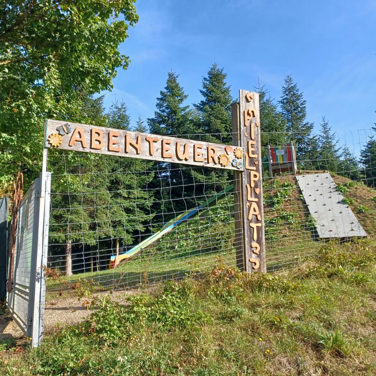 Ein Holzschild mit den Worten Abenteuer Spielplatz steht auf einem Holzzaun, umgeben von Bäumen und Grün. Dahinter befindet sich eine Rutsche und eine Kletterstruktur.