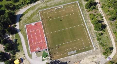 Eine Draufsicht auf einen Sportplatz mit einem roten Tennisplatz und einem grünen Fußballfeld, umgeben von Bäumen und Wegen.