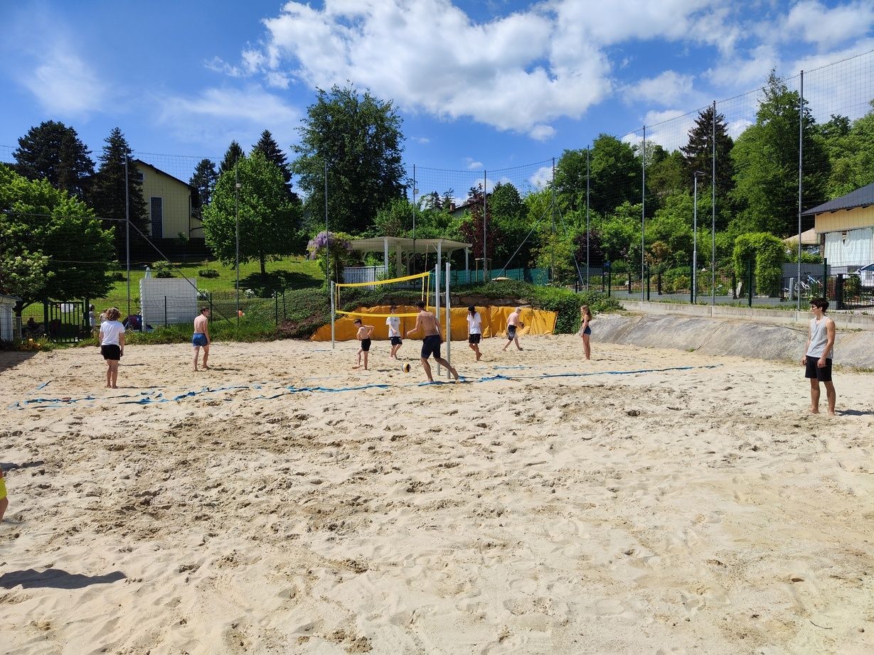 Eine Gruppe von Menschen spielt Beachvolleyball auf einem sandigen Feld mit Netz und Zaun. Bäume und ein Haus sind im Hintergrund.