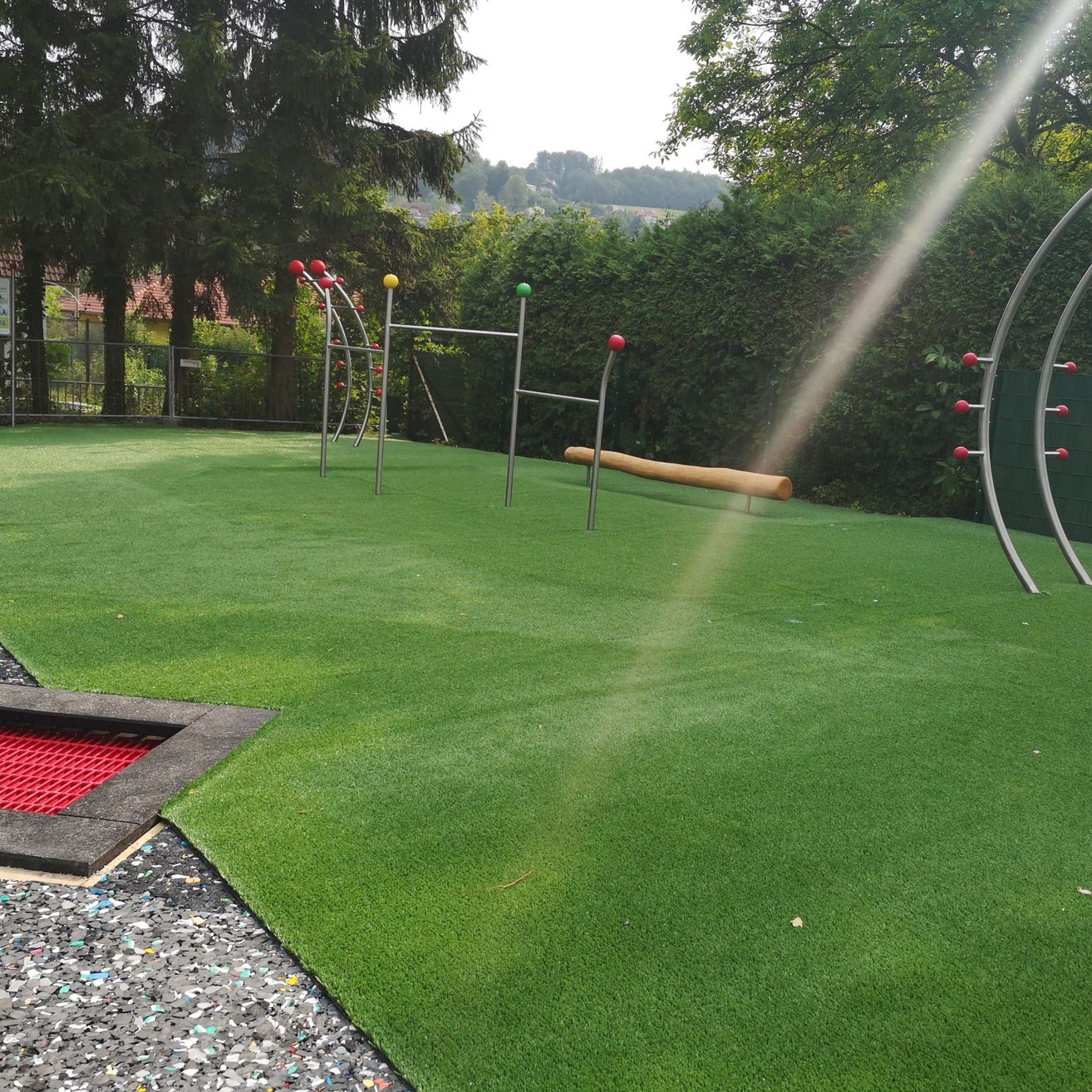 Ein Außenspielplatz mit Kunstrasen, Spielgeräten und einer Rutsche. Bäume und Büsche umgeben den Bereich, mit einem Zaun und Gebäude im Hintergrund.