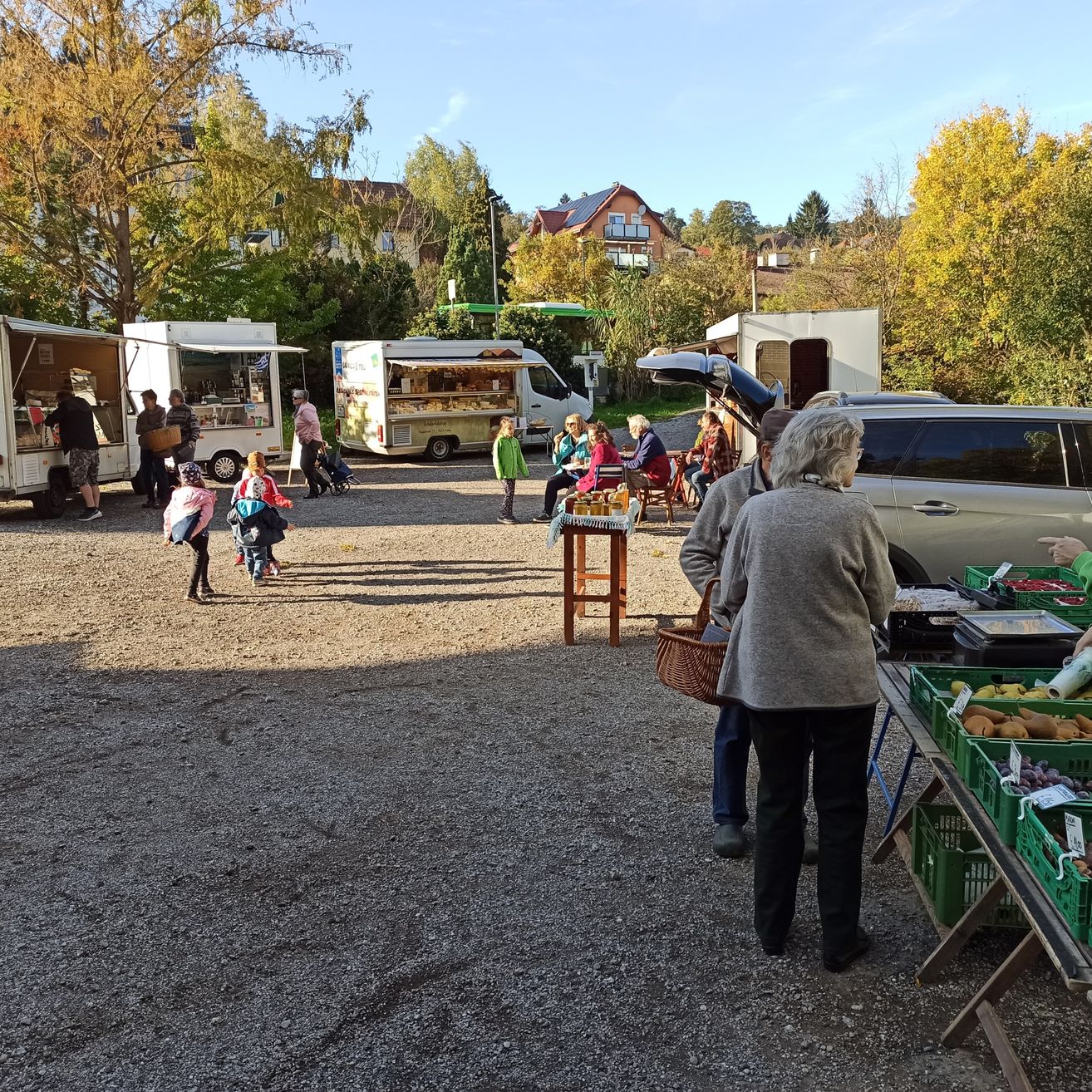 Ein geschäftiger Außenmarkt auf einem Schotterparkplatz mit mehreren Food-Trucks und Lieferwagen, bei dem Menschen die Stände durchstöbern, sich setzen und Körbe mit Obst und Gemüse tragen. Bäume und Gebäude sind im Hintergrund zu sehen.