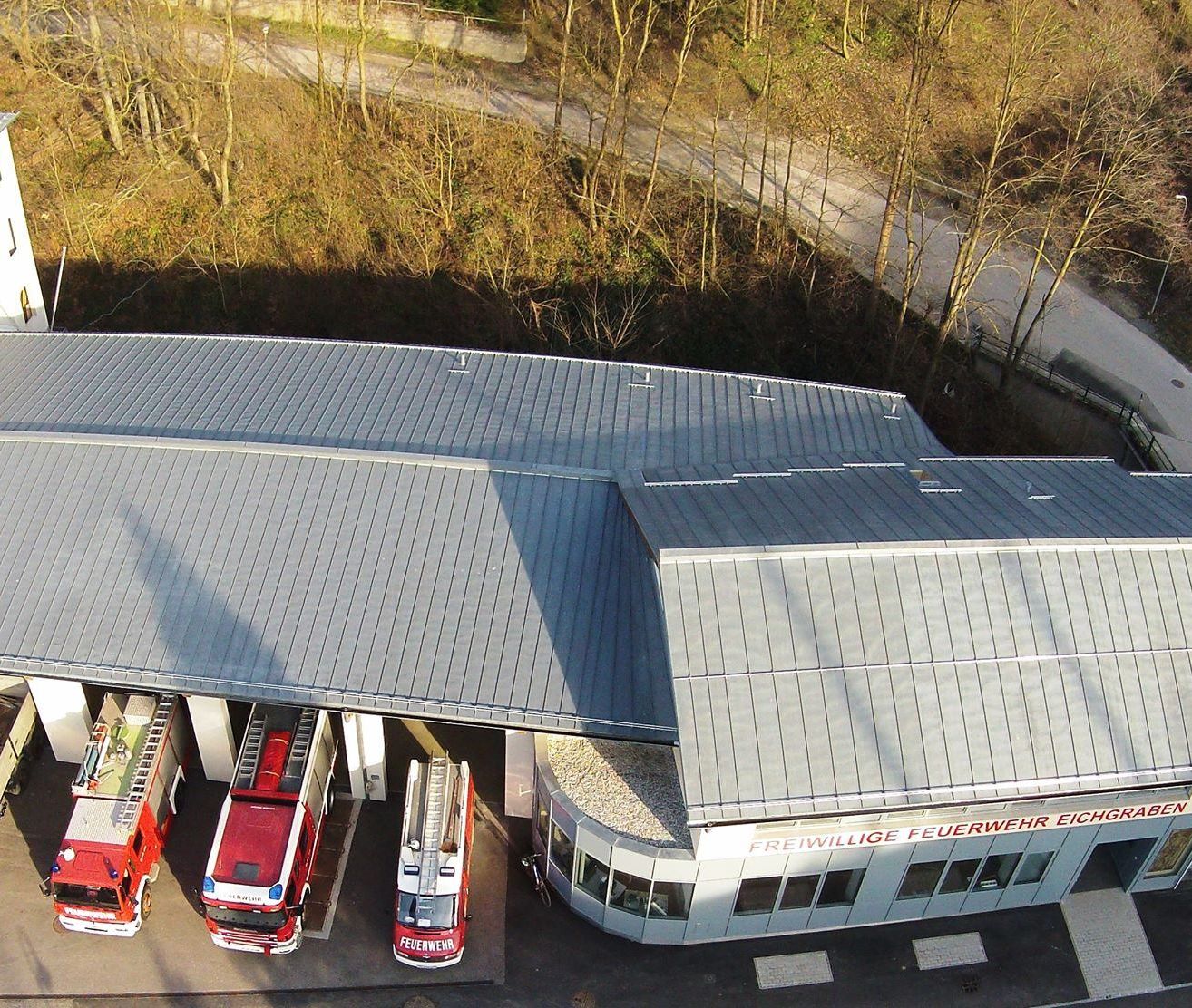 Ein Luftbild einer Feuerwehrwache mit grauen Dächern und roten Autos, die auf der Einfahrt geparkt sind. Der Text auf dem Gebäude lautet Freiwillige Feuerwehr Eich.