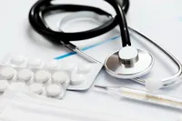 Ein Stethoskop, Thermometer und Pillen auf einer weißen Oberfläche. Das Stethoskop hat einen silbernen Schallkopf und schwarzes Schlauchmaterial. Es gibt zwei Pillenpackungen, eine mit drei Pillen.