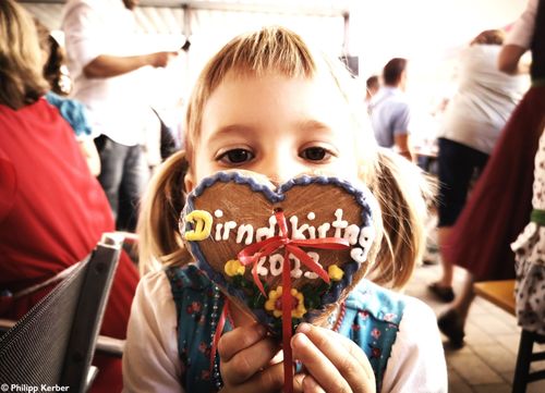 Ein kleines Mädchen hält einen herzförmigen Lebkuchen hoch, der mit bunter Zuckerguss und einer roten Schleife verziert ist und mit 'Dirndlkirtag 2013' beschriftet ist. Sie befindet sich inmitten einer Menschenmenge, wobei die Menschen und Objekte im Hintergrund verschwommen sind.