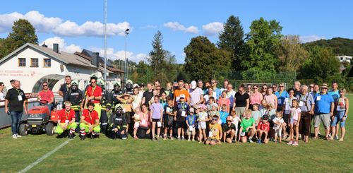 Eine große Gruppe von Menschen posiert für ein Foto auf einem Rasenfeld, einschließlich Feuerwehrleuten und Kindern, unter einem blauen Himmel mit Wolken und Bäumen.