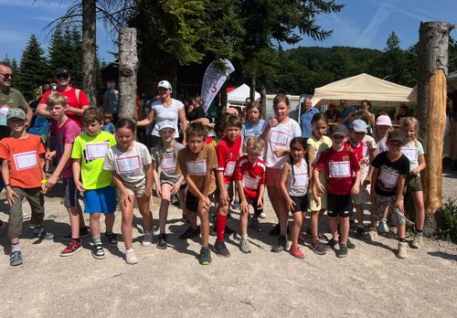 Gruppe von Kindern und Erwachsenen, die in einer Reihe mit nummerierten Startnummern in einem Waldgebiet stehen, mit Zelten und Bäumen im Hintergrund.