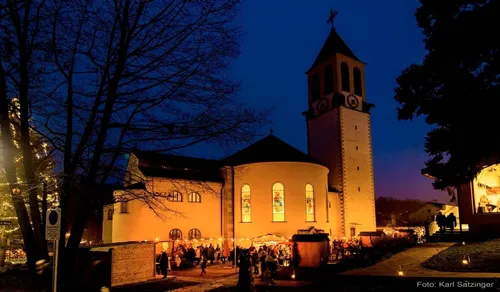 Bei Dämmerung ist eine Kirche mit einem hohen Turm und Buntglasfenstern beleuchtet und beherbergt eine geschäftige Veranstaltung im Freien mit Menschen, Zelten und einem Baum.