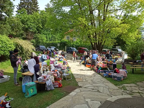 Ein Flohmarkt findet im Freien statt, viele Leute stöbern auf Tischen und dem Boden durch die Artikel. Ein Kleinkind geht auf einem Steinweg.