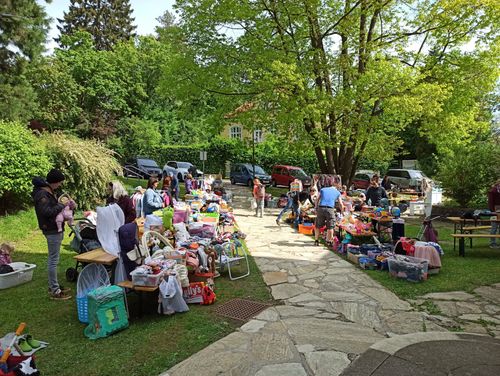 Ein Flohmarkt findet im Freien statt, viele Leute stöbern auf Tischen und dem Boden durch die Artikel. Ein Kleinkind geht auf einem Steinweg.
