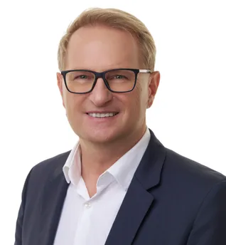 Ein Mann mit einer Brille und einem dunkelblauen Anzugsjackett mit weißem Hemd lächelt in die Kamera, vor einem schlichten weißen Hintergrund.