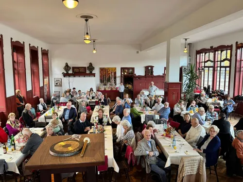 Ein Raum mit vielen älteren Menschen, die an Tischen sitzen, einige stehen, und einem großen runden Objekt auf einem Tisch.