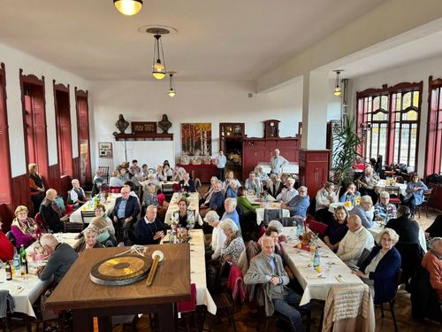 Ein Raum mit vielen älteren Menschen, die an Tischen sitzen, einige stehen, und einem großen runden Objekt auf einem Tisch.