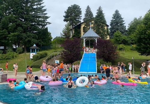 Eine Gruppe von Kindern und Erwachsenen schwimmt in einem Pool mit aufblasbaren Schwimmhilfen und einer blauen Wasserrutsche im Hintergrund.