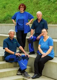 Vier Personen in blauen Hemden und Hosen, die auf Stufen mit einem Betonhintergrund und einem grün bewachsenen Hügel sitzen.