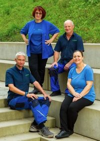 Vier Personen in blauen Hemden und Hosen, die auf Stufen mit einem Betonhintergrund und einem grün bewachsenen Hügel sitzen.