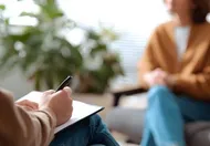 Eine Person schreibt in ein Notizbuch, während eine andere Person in einer Beratungssitzung zuhört.
