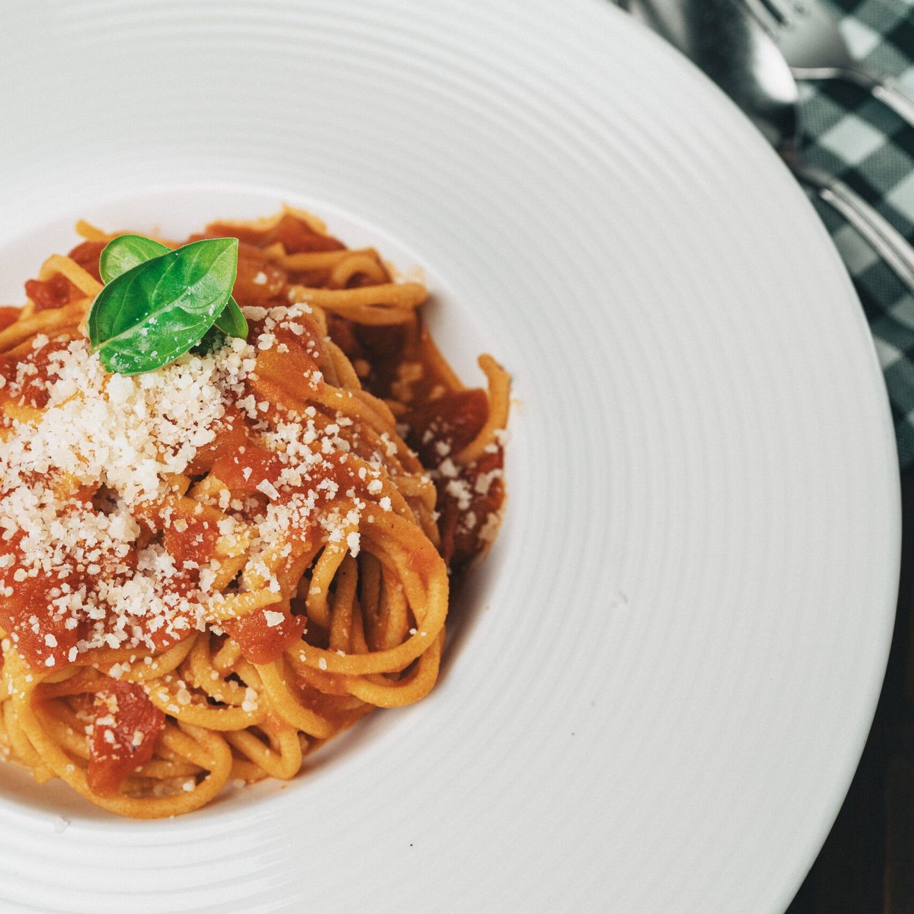 Ein weißer Teller mit Spaghetti und Tomatensoße, garniert mit Basilikumblättern und geriebenem Käse. Gabel und Löffel liegen daneben.