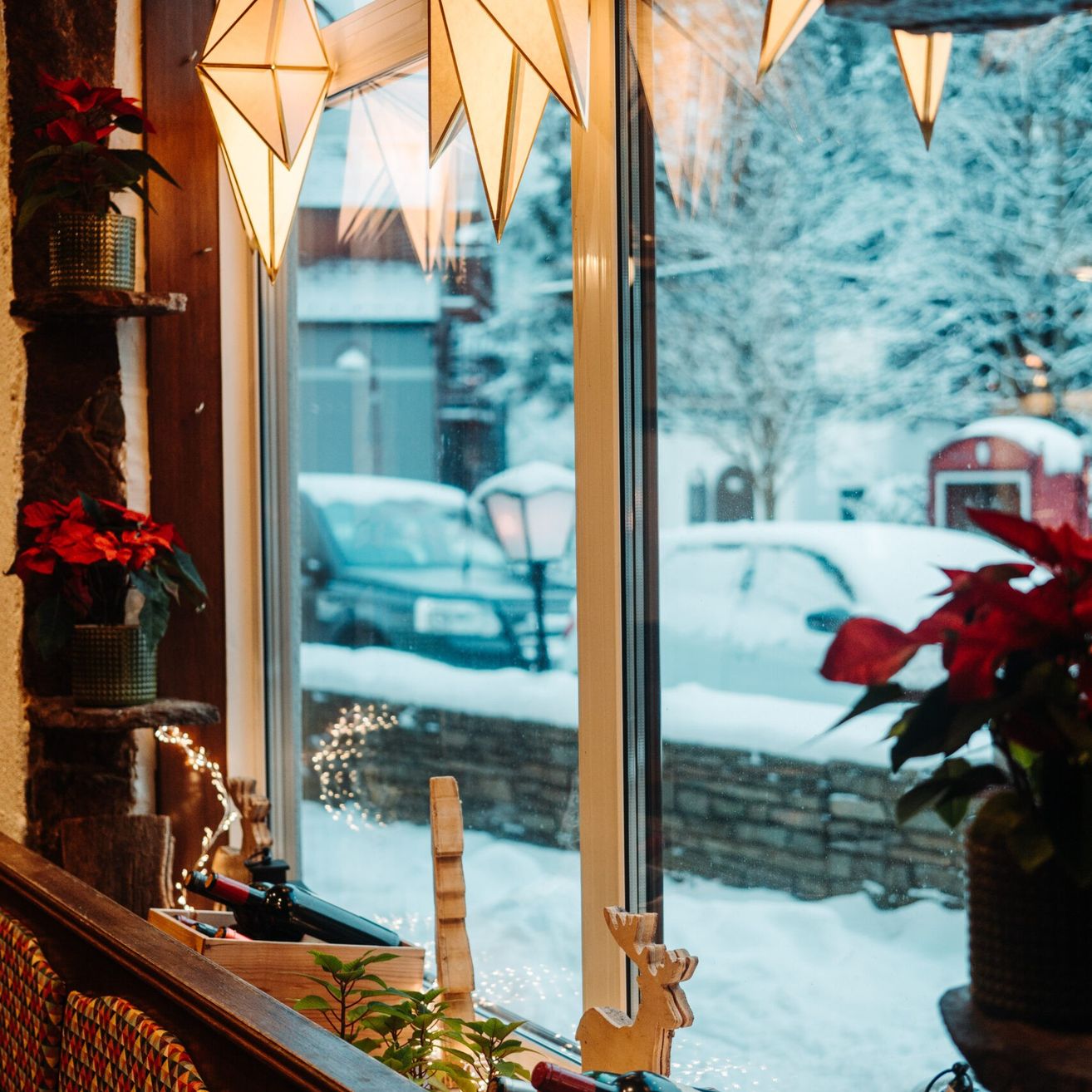 Eine gemütliche Innenansicht mit schneebedeckten Autos, Bäumen und einem Haus. Ein Fenster zeigt ein weihnachtliches Thema mit roten Blumen und Ornamenten.