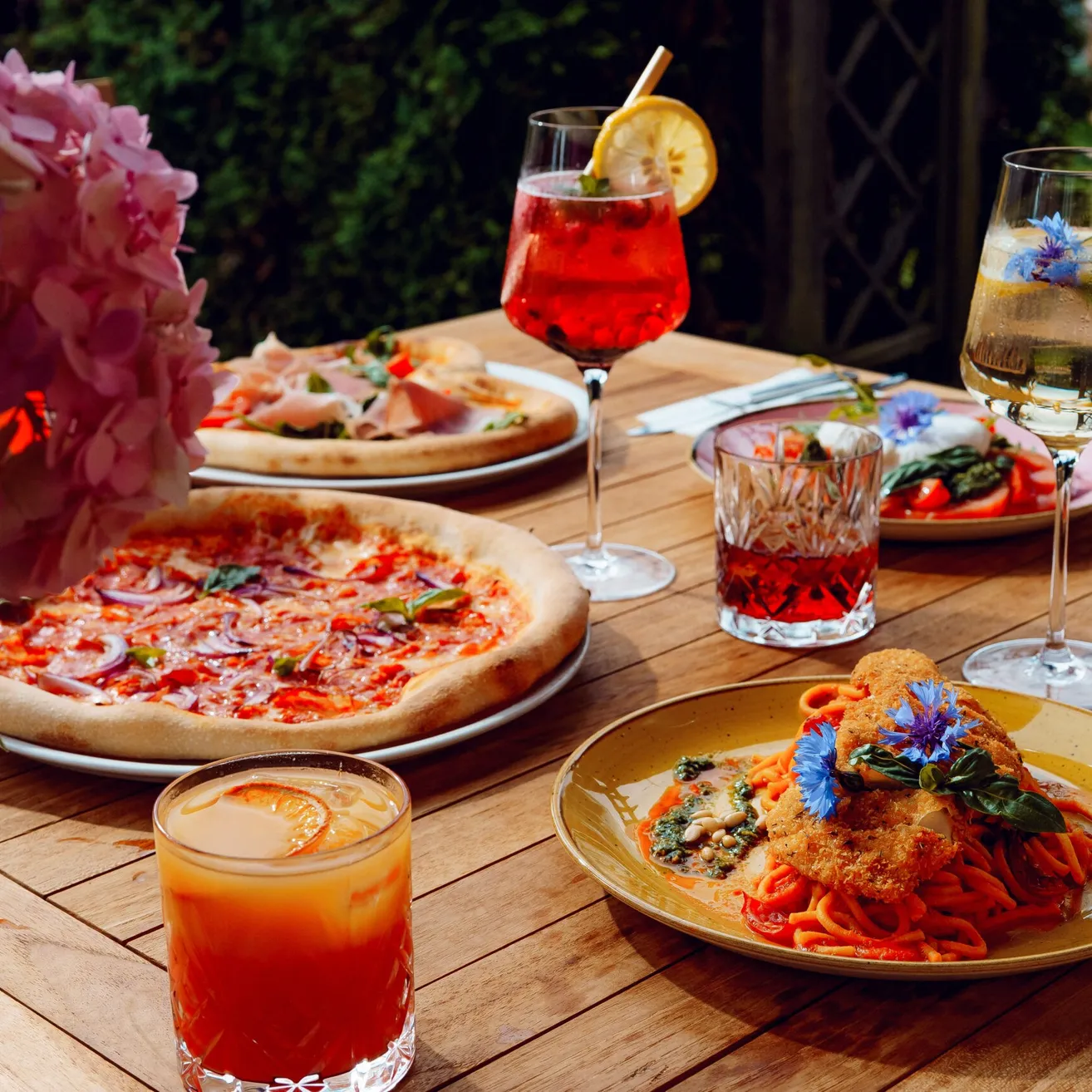 Ein Holztisch bietet verschiedene italienische Gerichte und Getränke an. Es gibt zwei Pizzen, einen Teller mit Nudeln und drei Gläser mit Getränken. Die Gerichte sind mit Blumen und Gemüse dekoriert.