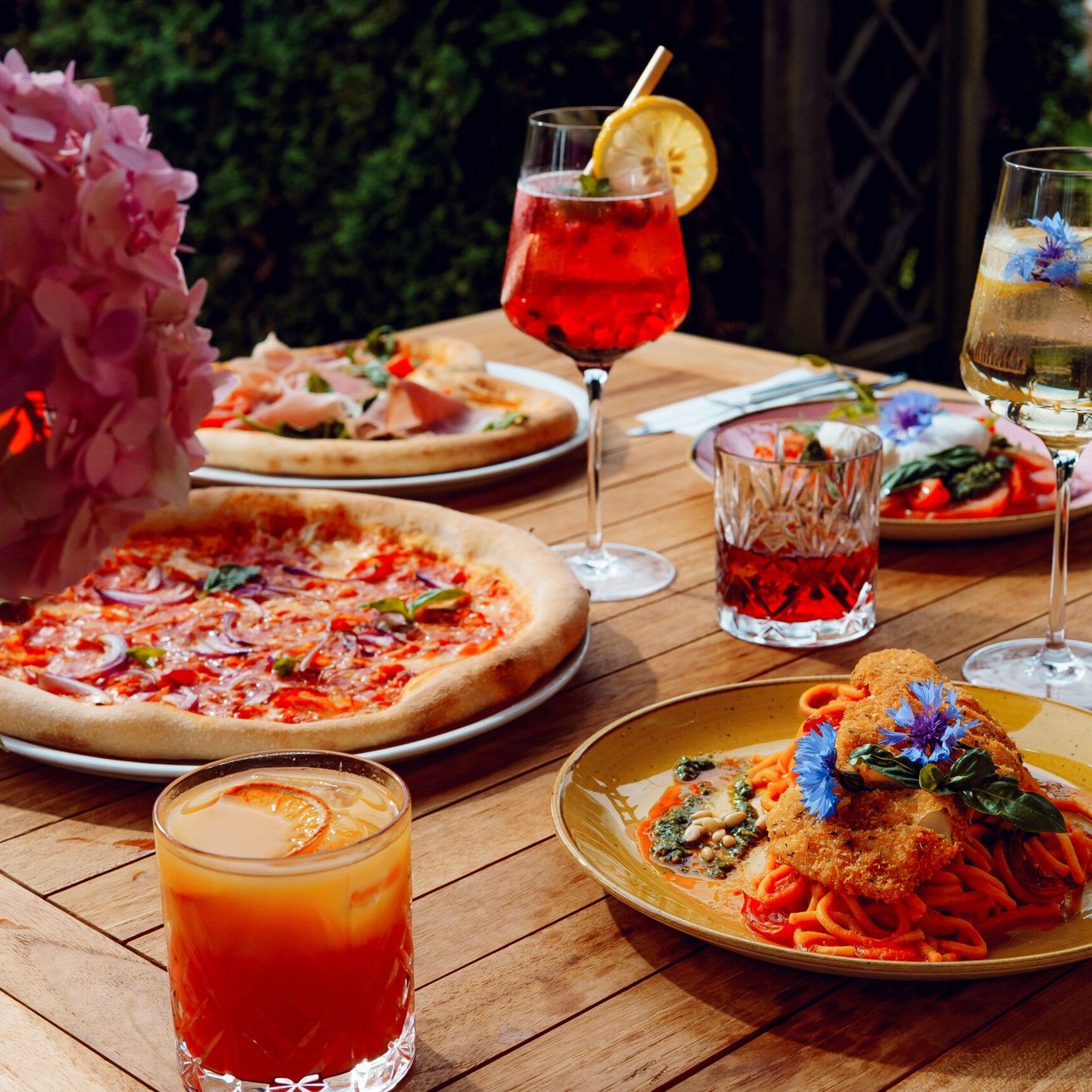 Ein Holztisch bietet verschiedene italienische Gerichte und Getränke an. Es gibt zwei Pizzen, einen Teller mit Nudeln und drei Gläser mit Getränken. Die Gerichte sind mit Blumen und Gemüse dekoriert.
