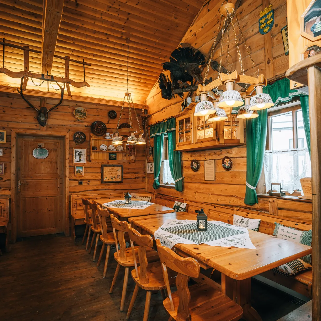 Ein rustikales Esszimmer mit Holzwänden und -böden. Zwei Holztische sind mit grünen Tüchern und Lampen gedeckt. Es gibt Geweihe und Tierköpfe an der Wand und eine Bank mit Kissen.