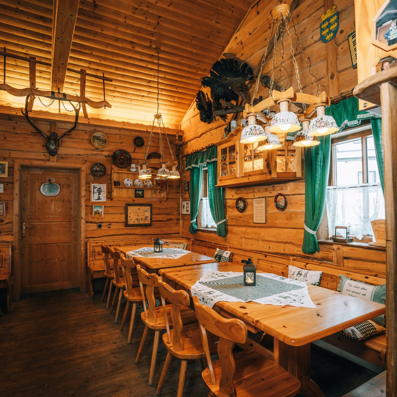 Ein rustikales Esszimmer mit Holzwänden und -böden. Zwei Holztische sind mit grünen Tüchern und Lampen gedeckt. Es gibt Geweihe und Tierköpfe an der Wand und eine Bank mit Kissen.