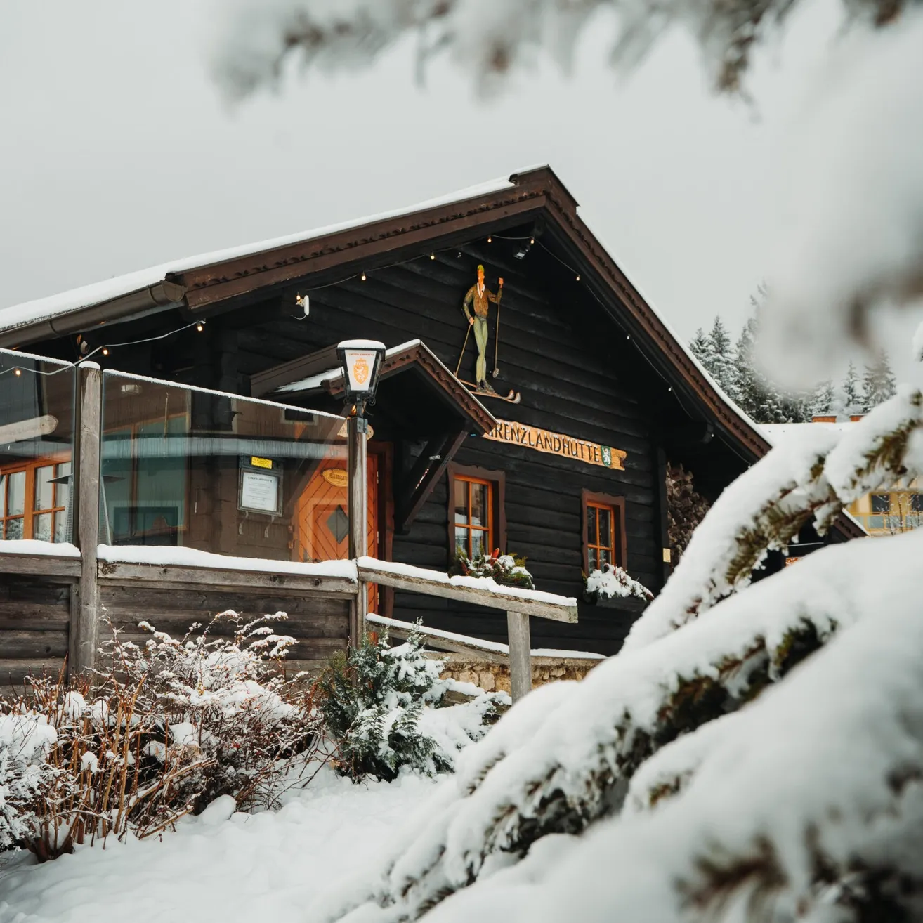 Eine verschneite Blockhütte mit einer Skifigur auf dem Dach, umgeben von schneebedeckten Bäumen und Pflanzen. Das Dach ist schneebedeckt.