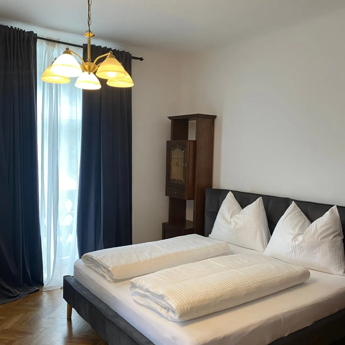 Ein ordentlich gemachtes Bett mit weißen Laken und Kissen in einem Zimmer mit einer Kronleuchter, Holzmöbeln und einem Fenster mit blauen Vorhängen.