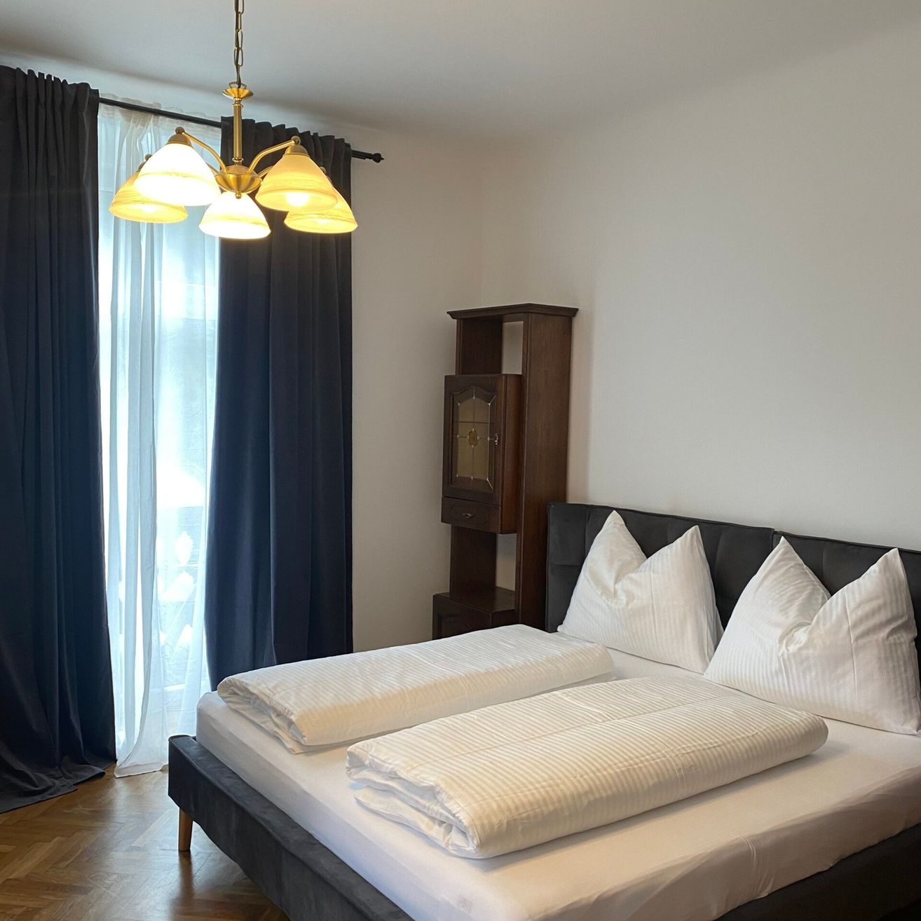 Ein ordentlich gemachtes Bett mit weißen Laken und Kissen in einem Zimmer mit einer Kronleuchter, Holzmöbeln und einem Fenster mit blauen Vorhängen.