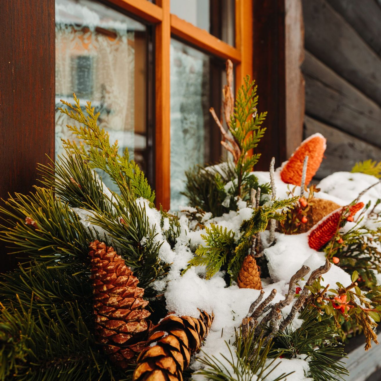 Ein Nahaufnahme eines Fenstersimses mit einer dekorativen Weihnachtsgirlande aus Tannenzapfen, Zweigen und roten Beeren, die mit Schnee bedeckt ist. Die Girlande steht an einer Holzwand.