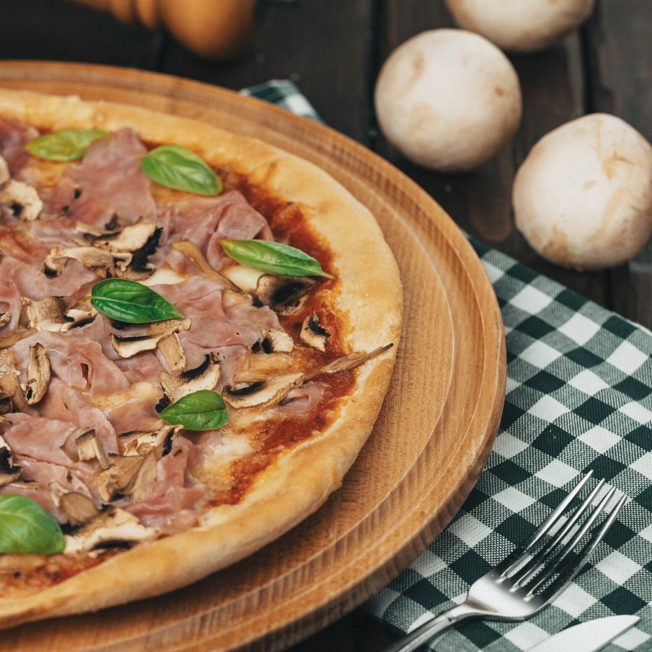 Eine Pizza mit Pilzen, Schinken und Basilikumblättern auf einem Holzteller, begleitet von einer Gabel und einem Messer auf einem grün-karierten Tuch.