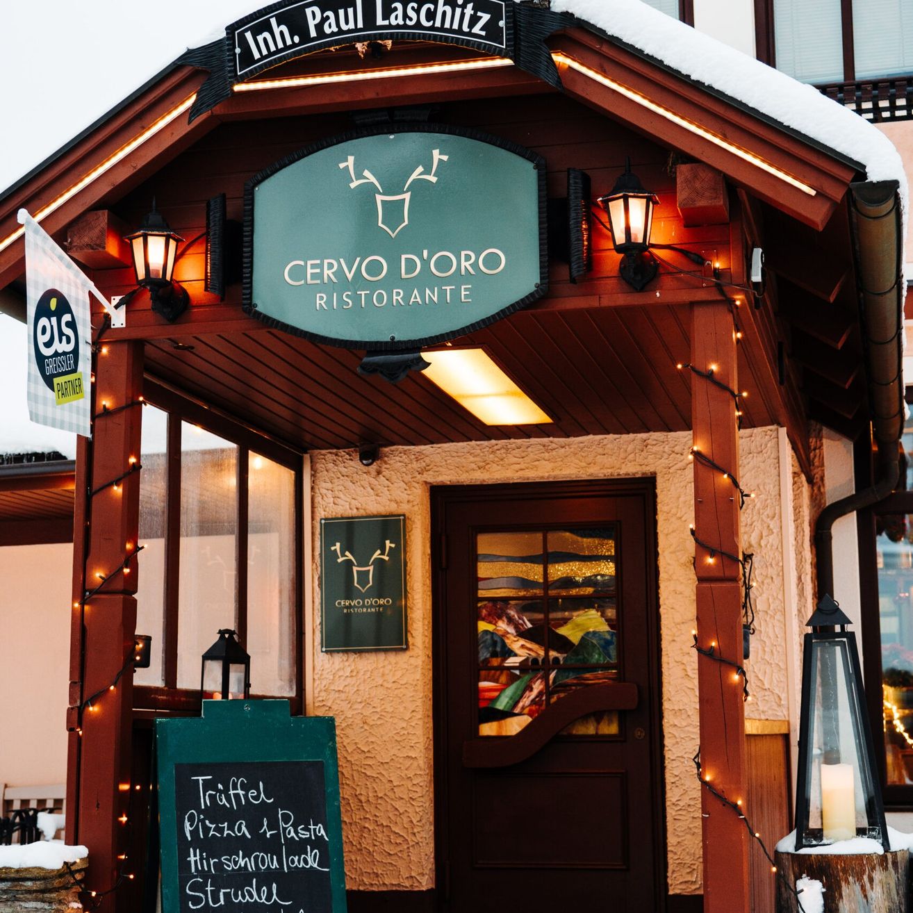 Der Eingang des Cervo D'Oro Ristorante, geschmückt mit einem grünen Schild und Weihnachtslichtern. Ein schwarzes Menübrett vor dem Eingang bietet Gerichte wie Trüffelpizza und Pasta an.