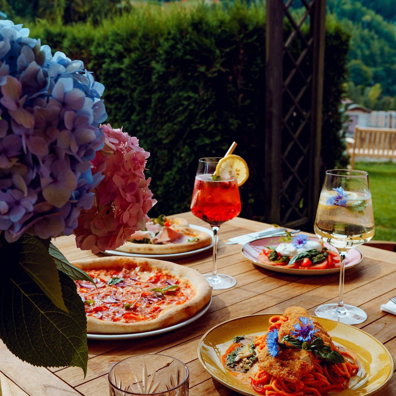 Ein hölzerner Tisch mit mehreren Tellern mit Essen, einem Glas Wein und einem Glas Wasser, vor dem Hintergrund von Blumen, einem Busch und einer Bank.