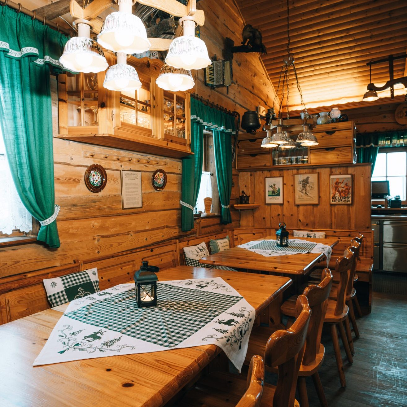 Ein rustikaler Essbereich mit Holztischen und Bänken, geschmückt mit grünen und weißen Tischdecken. Holzwände, Deckenlampen und Vorhänge vervollständigen die gemütliche Atmosphäre.