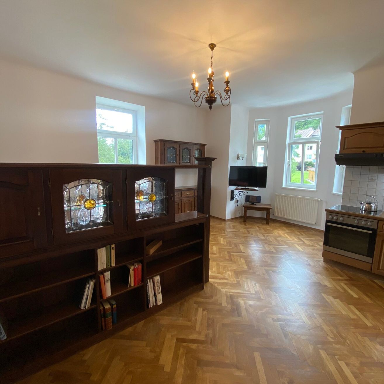 Wohnzimmer mit Holzböden, einer Kristallleuchte, einem Fernseher, einem Heizkörper und einer Küche mit Backofen.