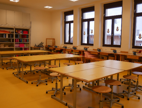 Ein leerer Klassenraum mit gelbem Boden und in Reihen angeordneten Tischen, mit Fenstern und Schränken im Hintergrund.