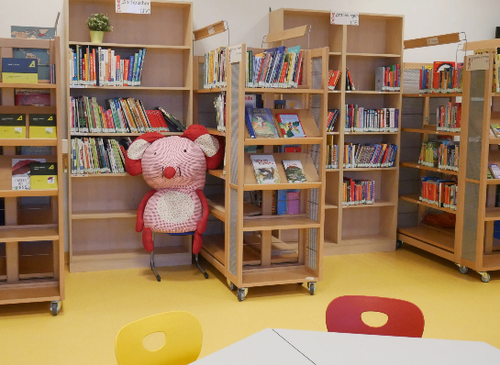 Eine Bibliothek mit Holzregalen voller Bücher, einem rosa Plüschtier auf einem Stuhl und einem gelben Boden.