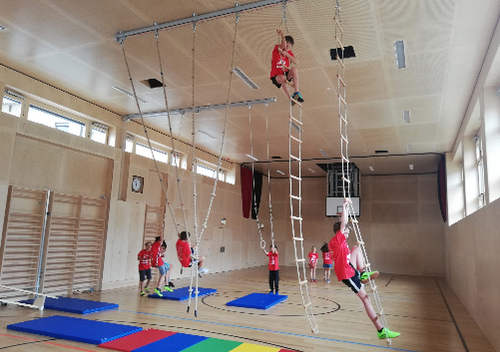 Eine Gruppe von Kindern in roten Shirts klettert in einer Turnhalle an Seilen. Sie hängen mit den Füßen an den Leitern an der Decke. Die Turnhalle hat Holzböden und blaue Matten auf dem Boden. Es gibt auch andere Kinder, die an der Seite stehen.