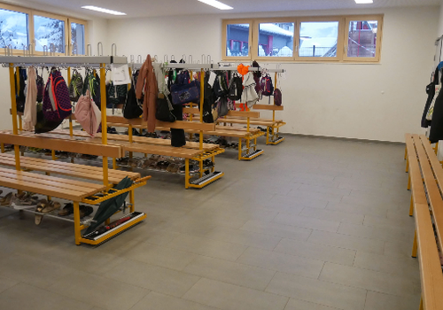 Ein sauberer, organisierter Raum mit Holzbänken und Gestellen, die verschiedene Taschen und Schuhe halten. Der Raum hat weiße Wände und Fenster.