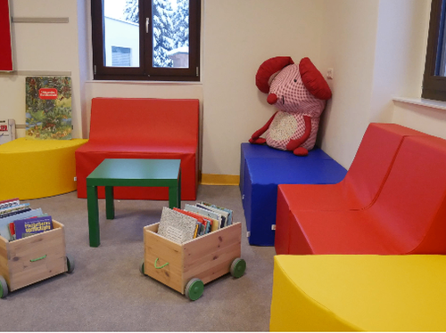 Ein Kinderspielzimmer mit bunten Möbeln, einem Plüschtier und Büchern auf einem Wagen. Ein Fenster zeigt eine verschneite Landschaft draußen.