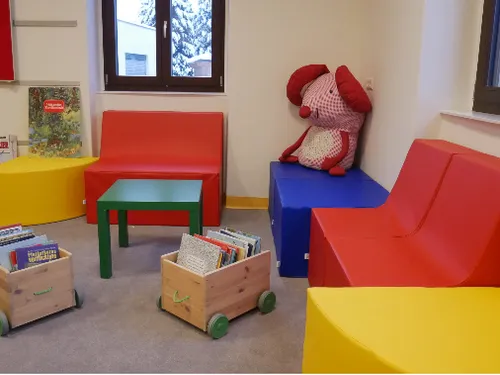 Ein Kinderspielzimmer mit bunten Möbeln, einem Plüschtier und Büchern auf einem Wagen. Ein Fenster zeigt eine verschneite Landschaft draußen.