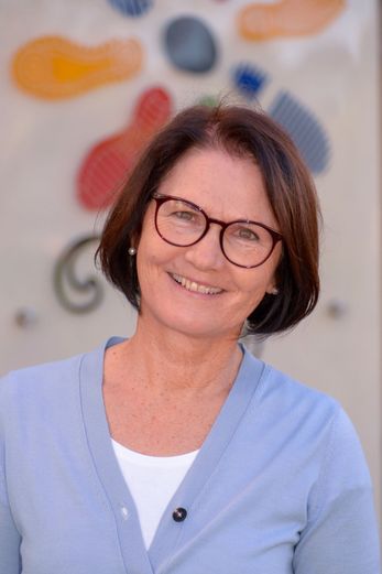 Eine Frau mit kurzen braunen Haaren und einer Brille lächelt in die Kamera. Sie trägt ein hellblaues Oberteil und steht vor einer gemusterten Wand.